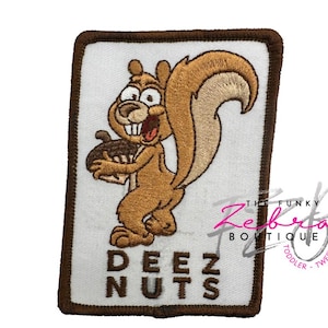 May include: Embroidered patch featuring a cartoon squirrel holding an acorn. The squirrel is brown with a large, fluffy tail and a surprised expression. The text "DEEZ NUTS" is below the squirrel, with a brown border.