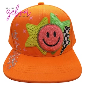 May include: Bright orange baseball cap with a smiley face patch, star and lightning bolt accents. The cap features a flat brim and is embellished with sequins and the text "THE FUNKY ZEBRA BOUTIQUE".