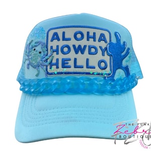 May include: Light blue trucker hat with the words "ALOHA HOWDY HELLO" in a rectangular patch. The hat features a blue crab and a blue wave design. The brim and mesh back are light blue.