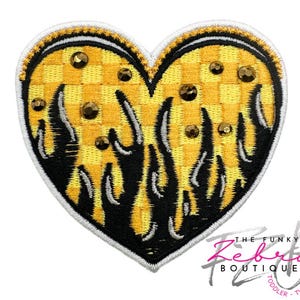May include: A heart-shaped patch with a yellow and black checkered pattern, embellished with brown rhinestones. Black flames emanate from the bottom of the heart. The patch has a white border and the text "THE FUNKY ZEBETA BOUTIQUE TODDLER TWEEN" in the bottom right corner.