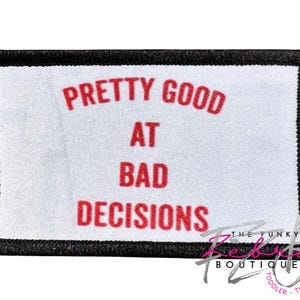 May include: A rectangular patch with a white background and a black border. The text "PRETTY GOOD AT BAD DECISIONS" is printed in red. The bottom right corner has a logo that says "THE FUNKY REBEL BOUTIQUE TODDLER TWEEN".