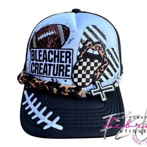 May include: A black and white baseball cap with a football design and the text "BLEACHER CREATURE". The cap features a leopard print brim, a cross pendant, and a football design on the front.