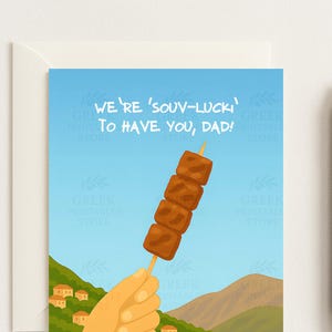 May include: Greeting card with a blue sky background and the text "WE'RE 'SOUV-LUCKI' TO HAVE YOU, DAD!". The card features an illustration of a hand holding a skewer of grilled meat. A black pen is on the right.