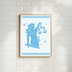 May include: A framed print featuring a light blue illustration of the Libra zodiac sign. The artwork depicts a woman holding scales and a sword, surrounded by stars. The print has a light wood frame and is displayed against a white brick wall.