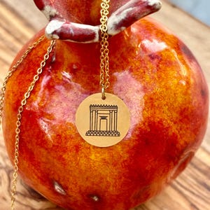 May include: A gold-colored necklace with a circular pendant featuring a black line drawing of a building with columns. The necklace hangs against a vibrant red and orange ceramic pomegranate.