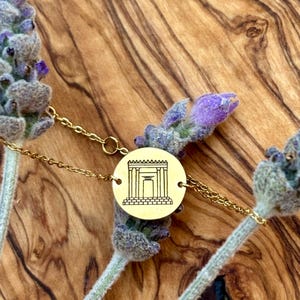 May include: A gold-colored bracelet featuring a circular pendant with a detailed architectural design. The bracelet is set against a backdrop of lavender sprigs and a wooden surface, creating a natural and elegant aesthetic.