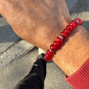 May include: A red beaded bracelet with silver accents. The bracelet is made of faceted, round beads and is worn on a wrist. The person is wearing a red long-sleeve shirt and black pants.