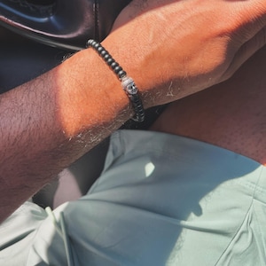 May include: A beaded bracelet featuring small, matte black spherical beads arranged in a continuous loop. The bracelet includes a single, central silver-toned metal charm shaped like a stylized human face with closed eyes and a serene expression.