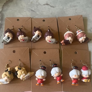 May include: A collection of handmade earrings featuring miniature cartoon figures. The earrings showcase various characters in different poses, with some holding objects. The figures are attached to gold-colored earring hooks and displayed on brown cardboard earring cards.