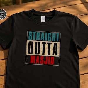 Puede incluir: Camiseta negra con el texto "STRAIGHT OUTTA MASJID" en un dise&ntilde;o rectangular. El texto es en blanco, verde azulado y rojo. La camiseta se muestra sobre una superficie de madera.