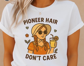 May include: White t-shirt with the text "PIONEER HAIR DON'T CARE" and a cartoon illustration of a woman wearing a hat and sunglasses, holding a drink with a lemon slice. The design includes floral and heart accents.