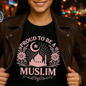 May include: Black t-shirt with the text "PROUD TO BE A MUSLIM" in pink, featuring a crescent moon, star, mosque silhouette, and floral accents. The person is wearing a brown leather jacket.