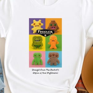 May include: A white t-shirt featuring a graphic print of six Fuggler monster plush toys in a grid. The text reads FUGGLER FUNNY UGLY MONSTER and Straight From The Dentist's Office of Your Nightmares. The monsters are yellow, brown, green, orange, and tan.