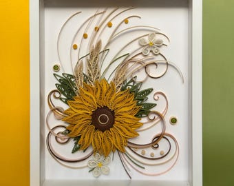 Puede incluir: Una obra de arte en quilling enmarcada que representa un girasol con pétalos amarillos y un centro marrón oscuro. La obra incluye hojas verdes, elementos similares al trigo y espirales decorativas, todo dentro de un marco blanco.