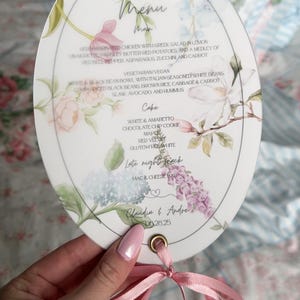 Może przedstawiać: Owalna karta menu weselnego z kwiatowymi ilustracjami akwarelowymi i różową wstążką. Menu zawiera opcje obiadowe i deserowe, w tym kurczaka, fasolę i ciasto. Karta jest trzymana w dłoni z różowym lakierem do paznokci.
