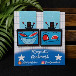 Könnte beinhalten: Zwei magnetische Lesezeichen mit fernsehköpfigen Charakteren, blauen Bildschirmen, roten Augen und Antennen. Die Trägerkarte ist weiß mit blauem Gittermuster, dem Text "Magnetic Bookmark" und den Social-Media-Konten von CoffinMothHollow.
