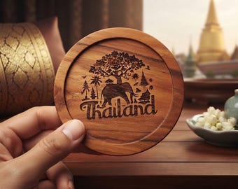 May include: A round wooden coaster featuring a laser-engraved design of an elephant standing beneath a large tree, surrounded by smaller trees and structures. The word Thailand is engraved in a stylised font at the bottom of the coaster surface.
