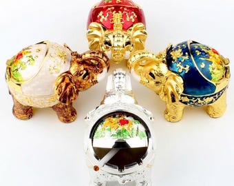 May include: Four elephant-shaped trinket boxes or ashtrays arranged in a circle. Three are gold-toned with red, white, and blue enamel lids featuring floral patterns. The fourth is silver-toned with a green and white landscape scene on the lid.