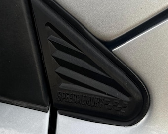 May include: A matte black plastic triangular car exterior trim piece featuring horizontal slats and the embossed text SPEEDMEMORY followed by a checkered flag graphic, mounted on the silver metallic body panel of a vehicle near the window.