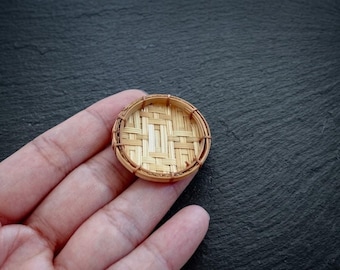 May include: A small, round, woven bamboo tray featuring a light tan colour and a detailed crisscross pattern. The tray has a thin, circular rim made of bent bamboo strips, showcasing a natural texture and a flat, shallow base design.