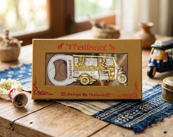 May include: A rectangular brown cardboard box with a clear window displaying a gold and silver metal bottle opener shaped like a tuk-tuk. The box features red text reading Thailand and Design By Thailand with small elephant icons on the top.