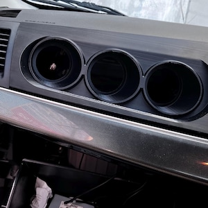 May include: A black automotive dashboard gauge pod featuring three circular openings for gauges. The unit is mounted above a carbon fiber textured trim piece. Below, an open glove compartment contains a small electronic box and a black case.