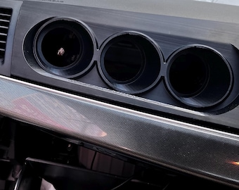 May include: A black automotive dashboard gauge pod featuring three circular openings for gauges. The unit is mounted above a carbon fibre textured trim piece. Below, an open glove compartment contains a small electronic box and a black case.