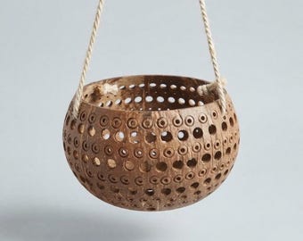 May include: A brown, spherical coconut shell hanging planter featuring a repeating pattern of circular cutouts of varying sizes. The bowl is suspended by two thick, light-coloured twisted jute ropes attached to the sides of the shell.