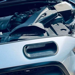 Op de afbeelding: Een zilverkleurige auto met een geopende motorkap die een zwarte kunststof luchtinlaatkanaal onthult achter een rechthoekige opening in de grille. De motoronderdelen zijn zichtbaar, inclusief een zwarte luchtinlaatschep naar het motorblok.