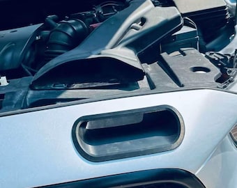 Könnte beinhalten: Ein silbernes Auto mit geöffneter Motorhaube, das einen schwarzen Kunststoff-Lufteinlasskanal hinter einer rechteckigen Kühlergrillöffnung zeigt. Die Motorkomponenten sind sichtbar, einschließlich eines schwarzen Lufteinlassschachts.