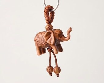 May include: A brown carved wooden elephant keyring featuring intricate heart and geometric patterns on its back. The elephant is suspended from a silver metal ring by a braided brown cord, with two additional small wooden beads hanging below.