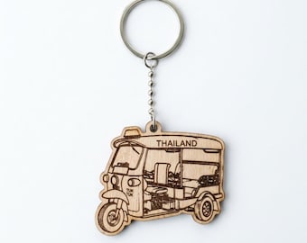 May include: A light brown wooden keyring featuring a laser-engraved illustration of a three-wheeled tuk-tuk vehicle. The design includes the word THAILAND printed on the roof and the text TUK TUK on the front panel, attached to a metal ring.