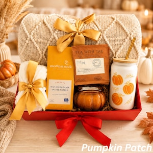 May include: A gift basket containing a beige textured throw blanket with a gold ribbon, a Moonstruck milk chocolate salted toffee bar, Tea With Tae earl grey tea, a small pumpkin-shaped candle, and a white glass cup with orange pumpkin prints.
