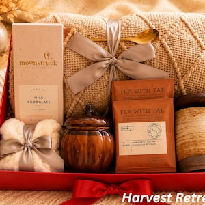 May include: A gift box containing a beige knit blanket tied with a grey ribbon and gold spoon, a Moonstruck milk chocolate bar, two Tea With Tae tea packets, a pumpkin-shaped candle, a white fuzzy item with a grey ribbon, and a ceramic mug.