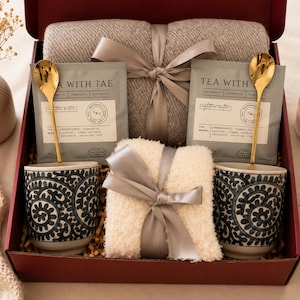 Puede incluir: Un juego de regalo en una caja burdeos incluye dos tazas estampadas, dos cucharas doradas, dos paquetes de té, una manta gris y una pequeña manta blanca, todo atado con cintas. También se ven una vela encendida y un jarrón con flores secas.