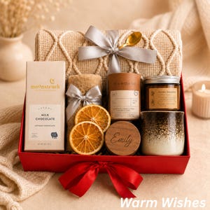 May include: A red gift box containing a beige knit blanket with a grey ribbon, a Moonstruck milk chocolate bar, a Tea With Tae canister, a candle labeled Aroma Sensation, a ceramic mug, two dried orange slices, and a wooden lid engraved with Emily.