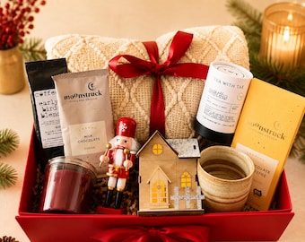 Könnte beinhalten: Eine rote Geschenkbox mit einer cremefarbenen, strukturierten Decke mit roter Schleife, einer Moonstruck-Milchschokolade, einer Tea With Tae-Dose, einer Nussknacker-Figur, einem Holzhaus-Ornament, einem Keramikbecher und einer dunkelroten Kerze.