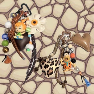 May include: Two keychains featuring cartoon characters. The first includes a Pebbles figurine, a brown heart, a white flower, and multi-colored beads. The second includes a Fred figurine, a leopard print heart, a tan heart, and multi-colored beads.