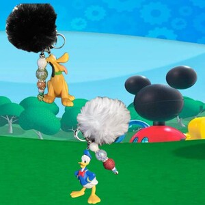May include: Two keychains featuring Disney characters. One has a black fluffy pom-pom and a Pluto figurine. The other has a white pom-pom and a Donald Duck figurine. Both have decorative beads and silver key rings.