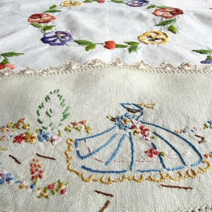 May include: A white linen tablecloth featuring hand-embroidered floral wreaths in yellow, purple, and red. The lower section displays an embroidered figure in a blue dress surrounded by small colourful flowers, finished with a cream crochet lace border.