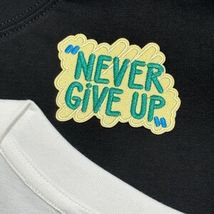 May include: Close-up of a black t-shirt with a yellow patch embroidered with the words "NEVER GIVE UP" in green. The patch has blue accents. A white t-shirt is visible in the lower portion of the image.