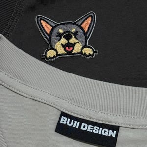 May include: Close-up of a gray t-shirt with a black label that reads "BUJI DESIGN." Above the label is a black and gray dog patch with orange ears and a smiling face. The dog patch is on a dark gray shirt.