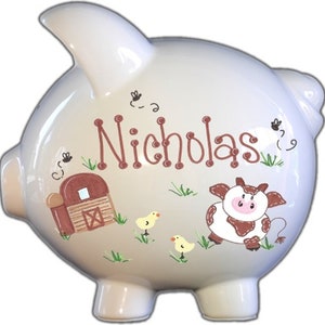 May include: White ceramic piggy bank with a brown barn, a brown and white cow, and two yellow chicks painted on it. The piggy bank is personalized with the name "Nicholas".