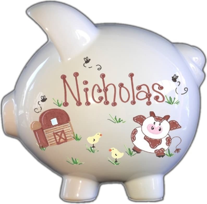 May include: White ceramic piggy bank with a brown barn, a brown and white cow, and two yellow chicks painted on it. The piggy bank is personalized with the name "Nicholas".