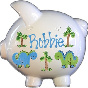 May include: White ceramic piggy bank with a blue and green dinosaur design, palm trees, and the name "Robbie" painted on the side.