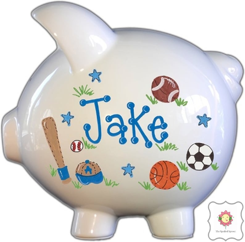 May include: White ceramic piggy bank with a blue and white baseball cap, baseball bat, baseball, basketball, football, and stars painted on it. The name "Jake" is written in blue letters on the side.