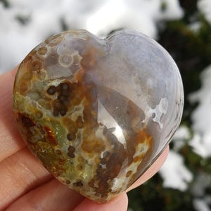 May include: A heart-shaped stone with a polished surface. The stone features a mix of colors, including brown, green, and white, with a translucent quality. The heart is held in a hand, with a snowy background.