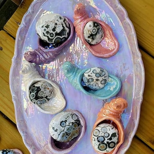 May include: An oval iridescent serving tray with a scalloped edge holds six small, shell-shaped ceramic dishes. Each dish contains a round, polished stone featuring a distinct black, white, and gray circular pattern, resembling an eye-like design.