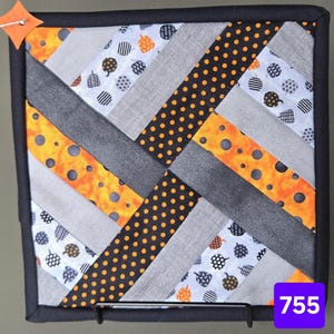 Pode incluir: Um pegador de panela quadrado de tecido com um design de patchwork diagonal em laranja, preto, branco e cinza. O padrão inclui bolinhas, faixas cinzas sólidas e um tecido branco com pequenos ícones de abóbora. Possui uma borda preta e uma pequena etiqueta laranja.