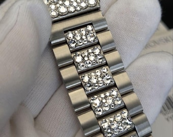 May include: A silver-toned metal watch band featuring a brushed finish and rectangular links. Each central link is embedded with a cluster of clear, sparkling round crystals that catch the light, creating a textured and reflective surface.